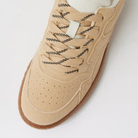 A close-up of a beige suede sneaker with black-and-white patterned laces, a white tongue label, and a brown gum sole, photographed on a light gray background.