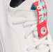 A close up of one of a pair of BALA Twelves in Flow White, which are bright white, athletic-style sneakers for nurses and healthcare workers. The photograph features the top of the shoe, including white laces, and a bright red ribbon that keeps the laces in place. The ribbon features a white and sky blue BALA logo, and fastens around the laces with a silver button, designed to look like a clock with the numeral 12 in the center.
