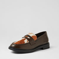 A single women's loafer shown in a side profile on a light gray studio background. The shoe features a dark brown leather upper with a contrasting front strap and a vamp panel made of brown and white cowhide.