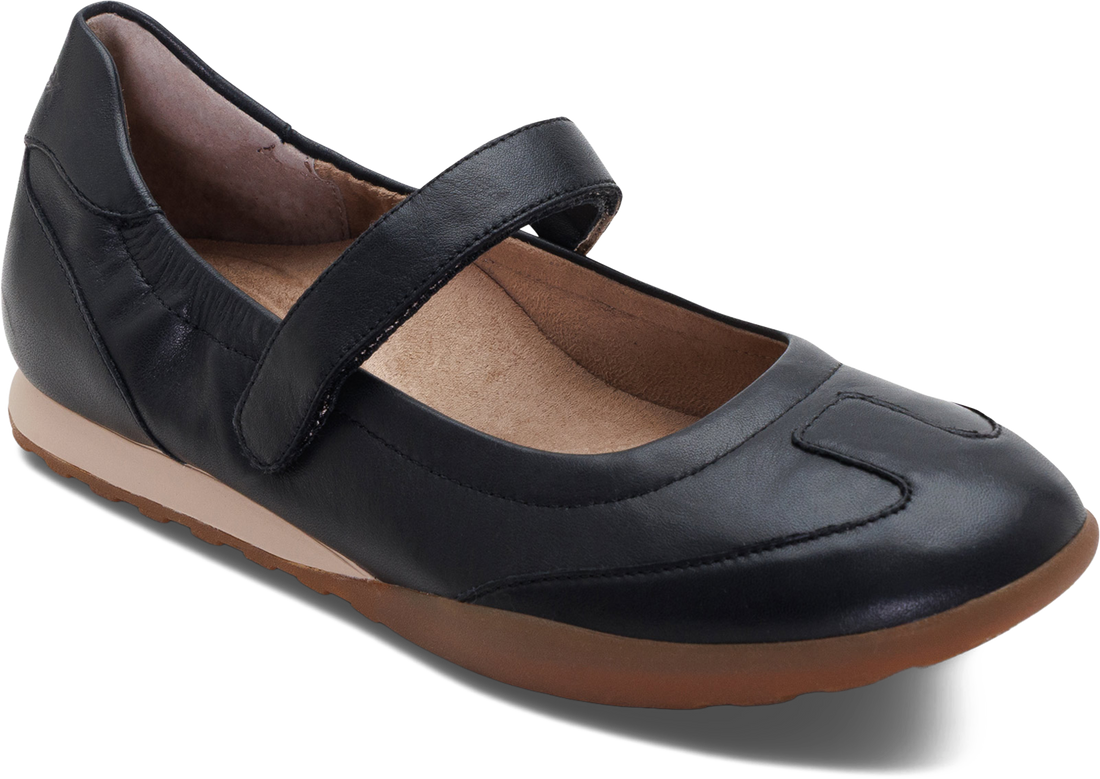 A black leather Mary Jane flat shoe with a hook-and-loop strap, brown sole, and tan interior lining, shown from a three-quarters angle on a white background.