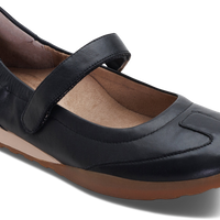 A black leather Mary Jane flat shoe with a hook-and-loop strap, brown sole, and tan interior lining, shown from a three-quarters angle on a white background.