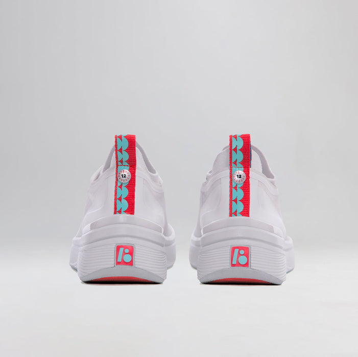 A pair of BALA Twelves in Flow White, which are bright white athletic-style sneakers for nurses and healthcare workers. They are photographed from the back; the heel features a bright red and sky blue ribbon with a pull tab, as well as a rubber BALA logo in the same colors. The ribbon is fastened with a silver button, designed to look like a clock, with the numeral 12 in the center.
