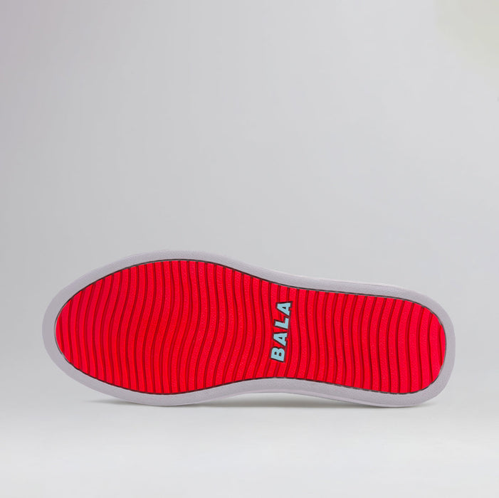 The sole of one of a pair of BALA Twelves in Flow White, which are bright white, athletic-style sneakers for nurses and healthcare workers. The sole is bright red with a white trim, a wavy tread, and a sky blue BALA logo. The photograph is taken against a solid white background.
